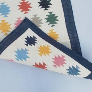 May include: A white rug with a blue border and colorful geometric patterns. The rug is folded in half, showing the underside of the rug.