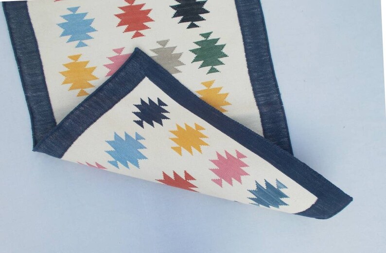 May include: A white rug with a blue border and colorful geometric patterns. The rug is folded in half, showing the underside of the rug.