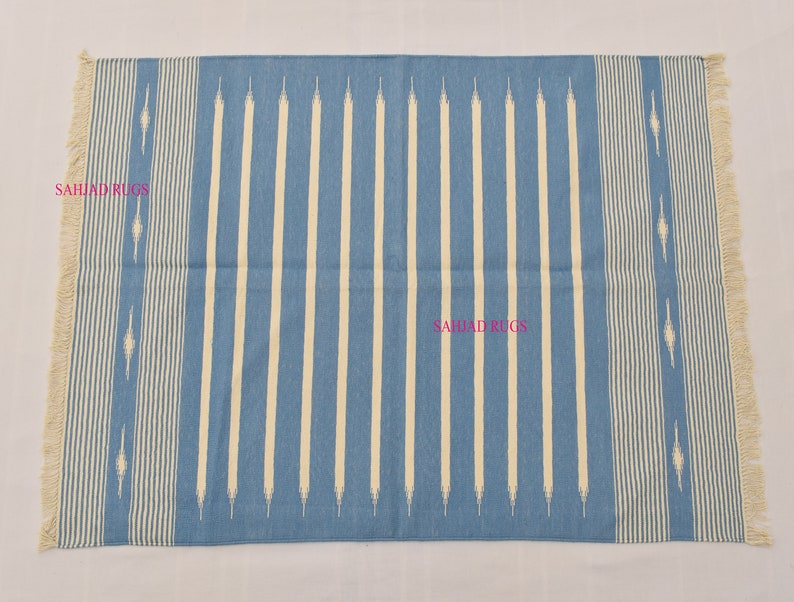 May include: A rectangular blue rug with vertical cream-colored stripes and fringed edges. The rug features a repeating pattern of cream-colored lines and decorative elements. The text "SAHJAD RUGS" is visible on the rug.
