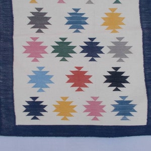 May include: A white rug with a blue border and a geometric pattern of colorful stars. The stars are in shades of blue, green, red, pink, yellow, and gray.