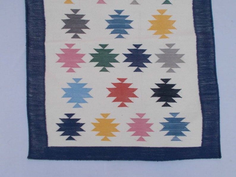 May include: A white rug with a blue border and a geometric pattern of colorful stars. The stars are in shades of blue, green, red, pink, yellow, and gray.
