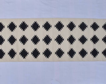 May include: A white runner rug with a black geometric pattern. The rug has a fringe on both ends.