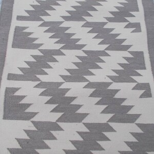 Op de afbeelding: Een wit en grijs geometrisch tapijt met een grijze rand. Het tapijt heeft een chevron-patroon.