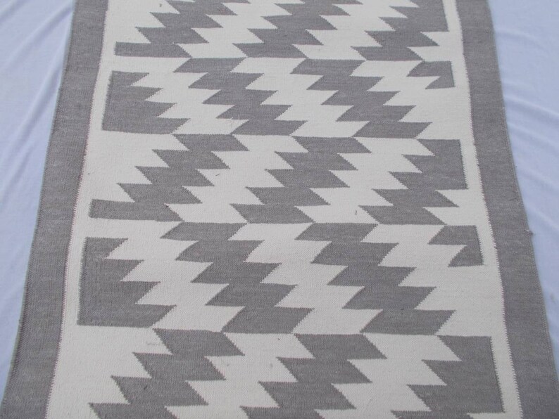 Op de afbeelding: Een wit en grijs geometrisch tapijt met een grijze rand. Het tapijt heeft een chevron-patroon.