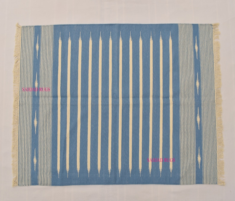 May include: A rectangular blue and white striped rug with fringed edges. The rug features vertical cream-colored stripes against a blue background. The design includes decorative elements and the text "SAHJAD RUGS".