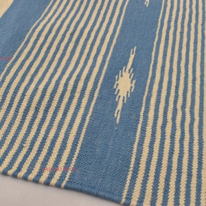 May include: A blue and cream-colored woven rug with vertical stripes and a fringed edge. The rug features a repeating pattern of alternating blue and cream-colored stripes, with a decorative diamond-like design.