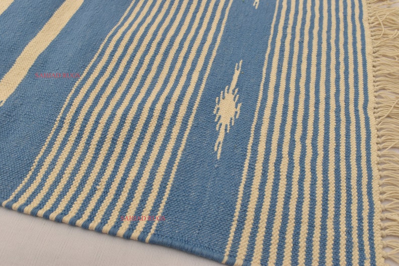 May include: A blue and cream-colored woven rug with vertical stripes and a fringed edge. The rug features a repeating pattern of alternating blue and cream-colored stripes, with a decorative diamond-like design.