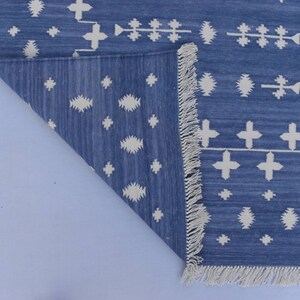 Puede incluir: Una alfombra azul y blanca con estampado y borde de flecos. La alfombra tiene un dise&ntilde;o geom&eacute;trico con cruces y diamantes blancos sobre un fondo azul.