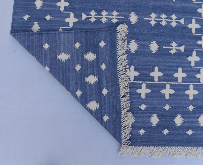 Puede incluir: Una alfombra azul y blanca con estampado y borde de flecos. La alfombra tiene un dise&ntilde;o geom&eacute;trico con cruces y diamantes blancos sobre un fondo azul.