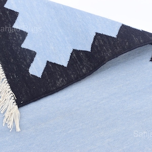 May include: A light blue rug with a black zigzag border and white fringe. The rug has a textured appearance and is folded over, revealing the underside. The text "Sahjad Rugs" is visible on the rug.