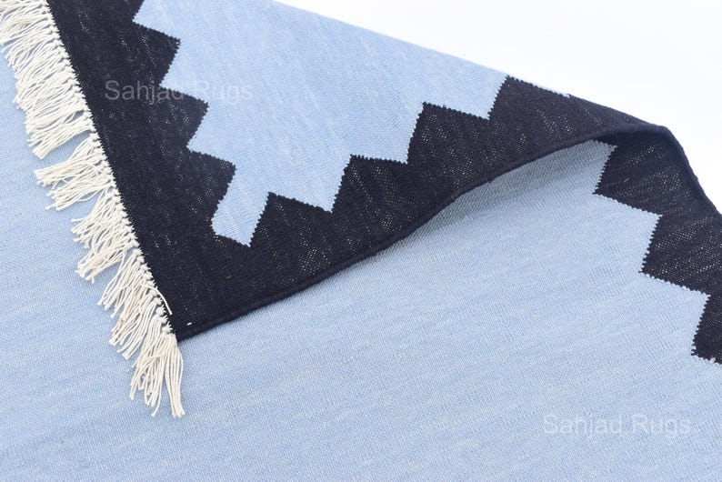 May include: A light blue rug with a black zigzag border and white fringe. The rug has a textured appearance and is folded over, revealing the underside. The text "Sahjad Rugs" is visible on the rug.