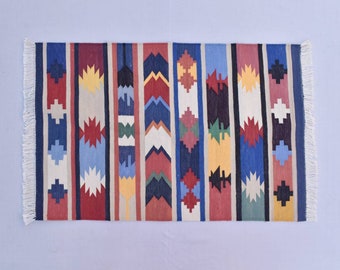 Puede incluir: Una alfombra rayada colorida con patrones geométricos en tonos de rojo, azul, amarillo, blanco y negro. La alfombra tiene un borde de flecos.