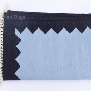 May include: A folded rectangular rug with a light blue center and a black border with a zig-zag design. The rug has a fringe on one end and the text "Sahjad Rugs" is visible. The rug is made of woven material.