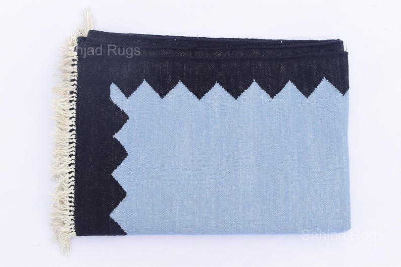 May include: A folded rectangular rug with a light blue center and a black border with a zig-zag design. The rug has a fringe on one end and the text "Sahjad Rugs" is visible. The rug is made of woven material.