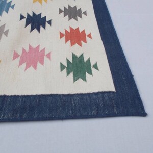 May include: A white rug with a blue border and colorful geometric patterns. The patterns are in shades of blue, green, yellow, pink, red, and gray.