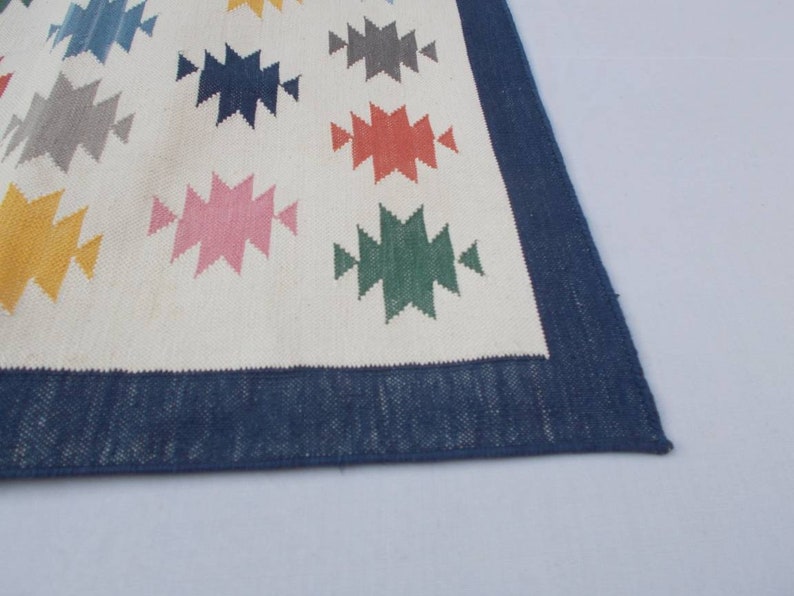 May include: A white rug with a blue border and colorful geometric patterns. The patterns are in shades of blue, green, yellow, pink, red, and gray.