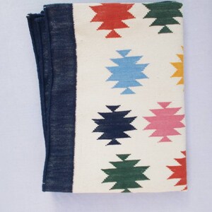 May include: A white woven rug with a navy blue border and colorful geometric patterns. The patterns are in shades of red, blue, green, pink, and yellow.