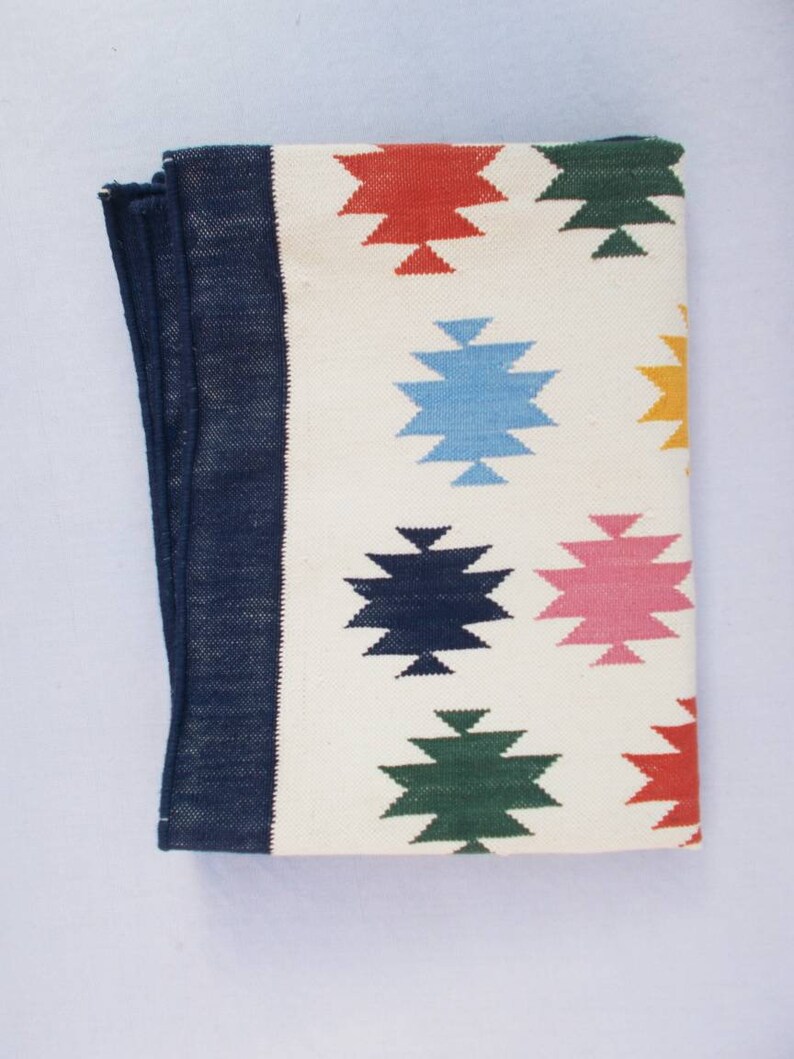 May include: A white woven rug with a navy blue border and colorful geometric patterns. The patterns are in shades of red, blue, green, pink, and yellow.