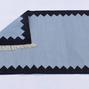 May include: A rectangular rug with a light blue center and a black zigzag border. The rug has a fringe on one end. The rug is made of a soft material and is suitable for home decor.