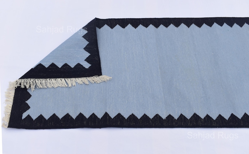 May include: A rectangular rug with a light blue center and a black zigzag border. The rug has a fringe on one end. The rug is made of a soft material and is suitable for home decor.