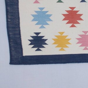 May include: A white rug with a blue border and a geometric pattern of colorful stars. The stars are in shades of blue, red, yellow, pink, and green.