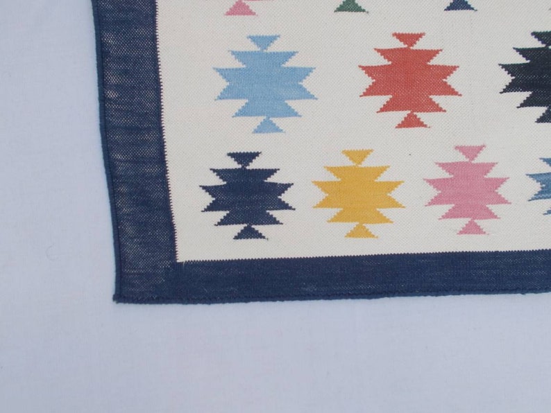 May include: A white rug with a blue border and a geometric pattern of colorful stars. The stars are in shades of blue, red, yellow, pink, and green.