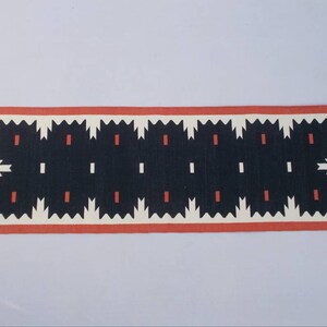 Puede incluir: Una alfombra geométrica de patrón negro y blanco con un borde rojo. La alfombra tiene un diseño de borde dentado.