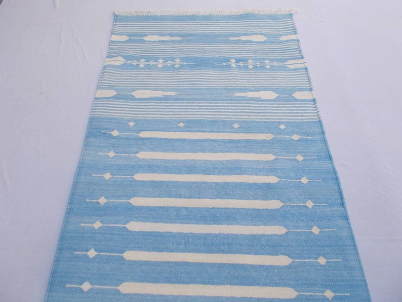 Puede incluir: Una alfombra de rayas azules y blancas con un patr&oacute;n geom&eacute;trico. La alfombra tiene un borde con flecos.