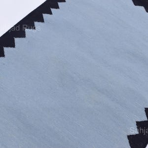May include: A rectangular rug with a light blue center and black zigzag borders. The rug has a simple design and a neutral color palette, suitable for various interior styles. The rug's dimensions are not visible.