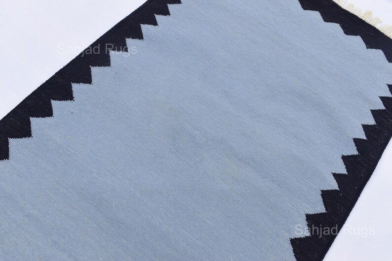 May include: A rectangular rug with a light blue center and black zigzag borders. The rug has a simple design and a neutral color palette, suitable for various interior styles. The rug's dimensions are not visible.