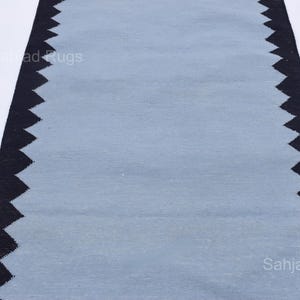 May include: A rectangular rug with a light blue center and a black zigzag border. The rug is made of a woven material and is displayed on a white background. The rug is a home decor item.