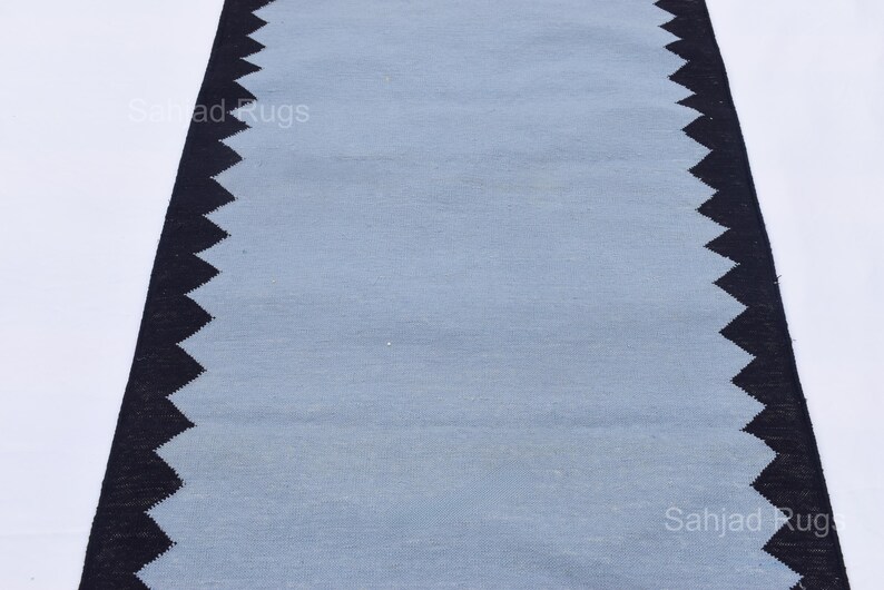 May include: A rectangular rug with a light blue center and a black zigzag border. The rug is made of a woven material and is displayed on a white background. The rug is a home decor item.