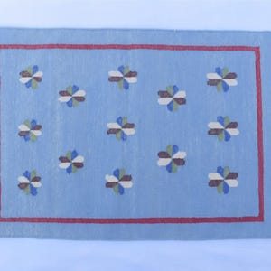 Op de afbeelding: Een rechthoekig, lichtblauw tapijt met een rode rand en crèmekleurige franjes. Het tapijt heeft een herhalend patroon van gestileerde bloemmotieven in tinten bruin, groen, blauw en crème. Het tapijt is waarschijnlijk gemaakt van geweven stof.