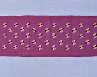 Op de afbeelding: Een lang, rechthoekig roze kleed met een herhalend patroon van kleine, veelkleurige bloemen. Het kleed heeft een dunne oranje rand en witte franjes aan de uiteinden.