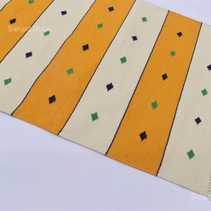 Puede incluir: Una alfombra rectangular con rayas diagonales amarillas y blanquecinas. La alfombra presenta peque&ntilde;os acentos en forma de diamante en verde y azul marino. La alfombra tiene un borde con flecos en un lado. La alfombra es de material tejido.