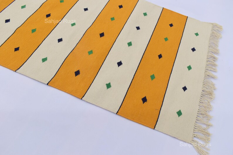 Puede incluir: Una alfombra rectangular con rayas diagonales amarillas y blanquecinas. La alfombra presenta peque&ntilde;os acentos en forma de diamante en verde y azul marino. La alfombra tiene un borde con flecos en un lado. La alfombra es de material tejido.