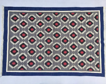 May include: A gray rug with a geometric pattern in black, red, and white. The rug has a blue border and fringe.
