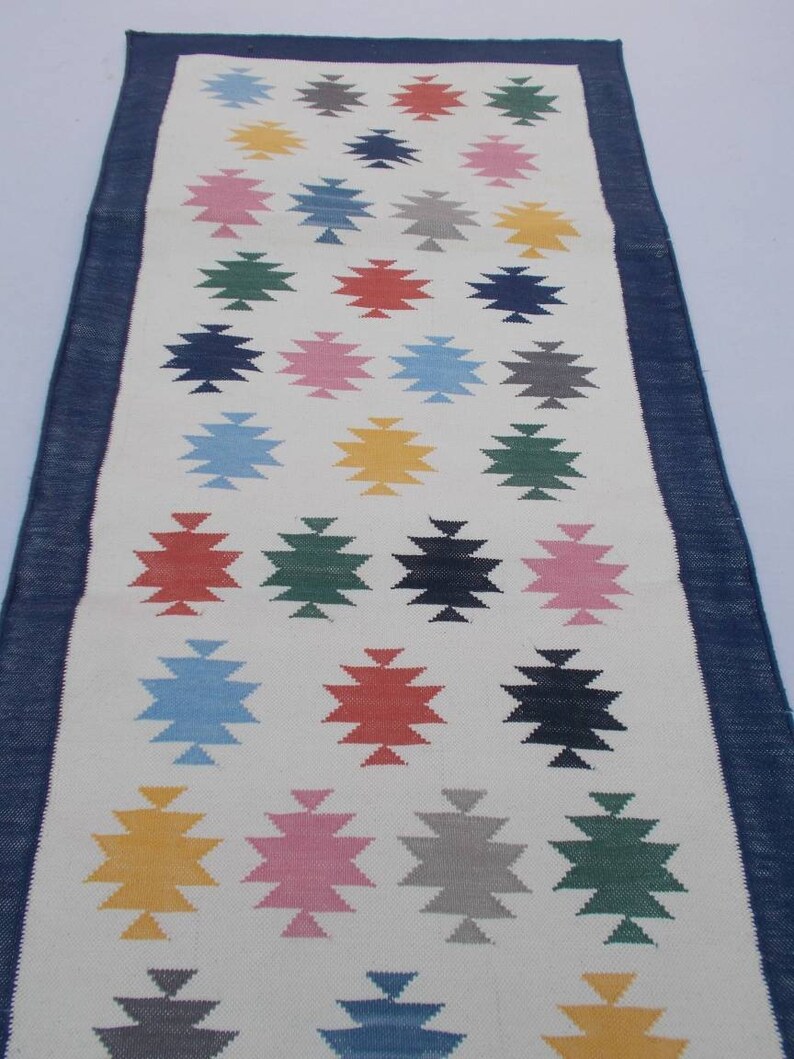May include: A white runner rug with a navy blue border. The rug features a repeating pattern of colorful geometric stars.