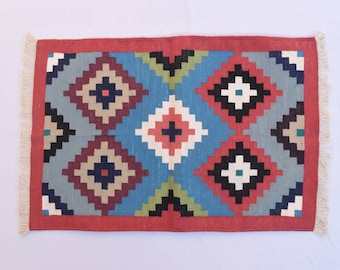 Puede incluir: Una alfombra geométrica colorida con un borde rojo y flecos blancos. La alfombra presenta un patrón repetitivo de diamantes en tonos de azul, verde, rojo, negro y blanco.