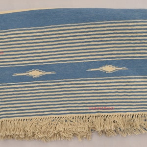 May include: Blue and cream-colored woven textile with horizontal stripes and geometric diamond-like shapes. The textile has a fringed edge. The design is a combination of blue and cream colors.
