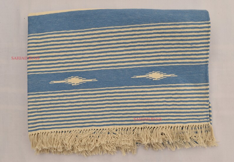 May include: Blue and cream-colored woven textile with horizontal stripes and geometric diamond-like shapes. The textile has a fringed edge. The design is a combination of blue and cream colors.
