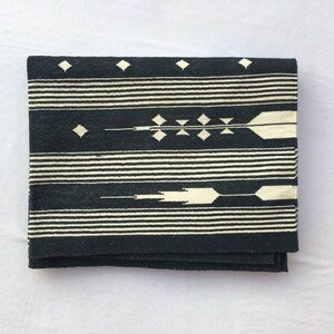 K&ouml;nnte beinhalten: Gefaltetes schwarz-wei&szlig; gewebtes Textil mit geometrischem Muster. Das Design zeigt horizontale Streifen, Rauten und pfeilartige Motive. Das Textil ist gefaltet und zeigt seine Textur und sein Design.