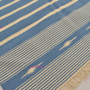 May include: Blue and cream-colored woven rug with a striped pattern. The rug features alternating stripes of light blue and cream, with a fringe along one edge. Decorative diamond shapes are incorporated into the design.