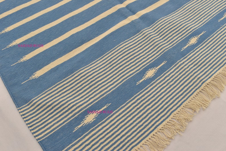 May include: Blue and cream-colored woven rug with a striped pattern. The rug features alternating stripes of light blue and cream, with a fringe along one edge. Decorative diamond shapes are incorporated into the design.