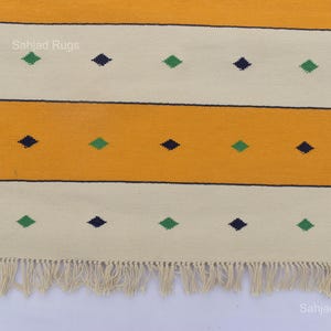 Puede incluir: Una alfombra rectangular con un patr&oacute;n de rayas amarillas y crema. La alfombra presenta detalles en forma de diamante azul oscuro y verde. La alfombra tiene flecos en la parte inferior. Las palabras "Sahjad Rugs" est&aacute;n impresas en la alfombra.