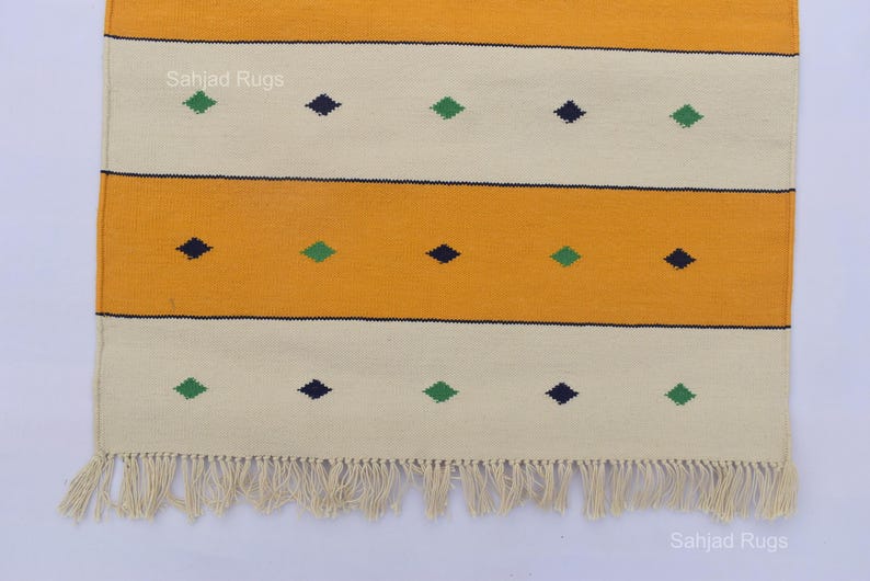 Puede incluir: Una alfombra rectangular con un patr&oacute;n de rayas amarillas y crema. La alfombra presenta detalles en forma de diamante azul oscuro y verde. La alfombra tiene flecos en la parte inferior. Las palabras "Sahjad Rugs" est&aacute;n impresas en la alfombra.