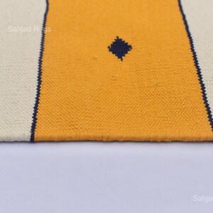 Puede incluir: Primer plano de una alfombra tejida con fondo crema, una franja central amarilla y un rombo azul oscuro. La alfombra tiene detalles de rombos verdes y bordes azul oscuro. La textura es visible.