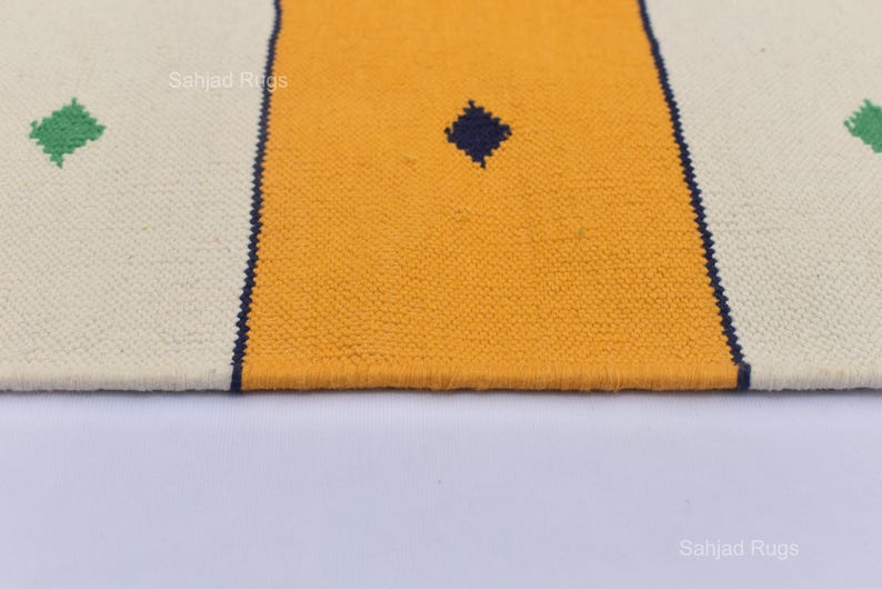 Puede incluir: Primer plano de una alfombra tejida con fondo crema, una franja central amarilla y un rombo azul oscuro. La alfombra tiene detalles de rombos verdes y bordes azul oscuro. La textura es visible.