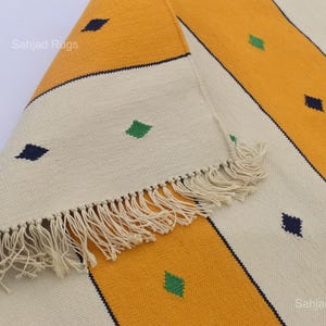 Puede incluir: Primer plano de una alfombra a rayas amarillas, crema y azul marino con flecos. La alfombra tiene un patr&oacute;n repetido de formas de diamante en verde y azul marino. La alfombra est&aacute; doblada, mostrando los flecos.