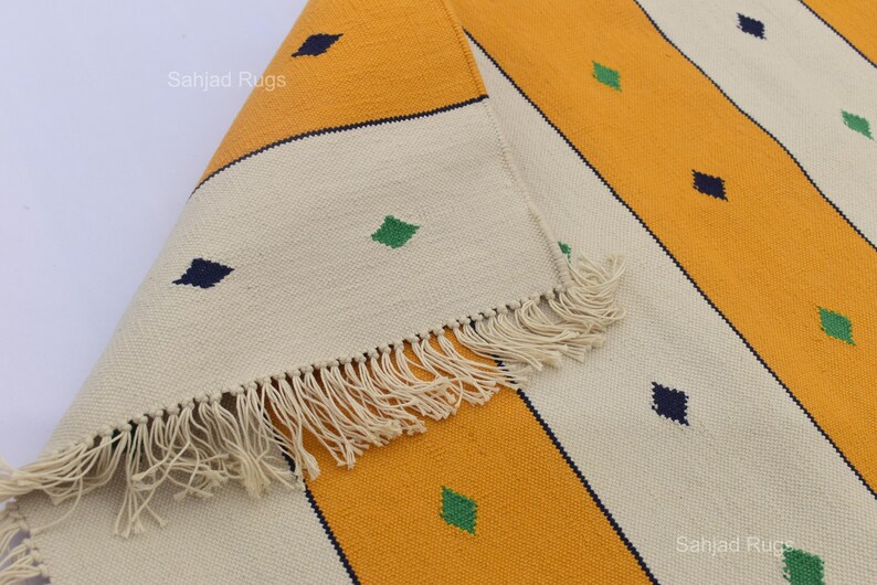 Puede incluir: Primer plano de una alfombra a rayas amarillas, crema y azul marino con flecos. La alfombra tiene un patr&oacute;n repetido de formas de diamante en verde y azul marino. La alfombra est&aacute; doblada, mostrando los flecos.